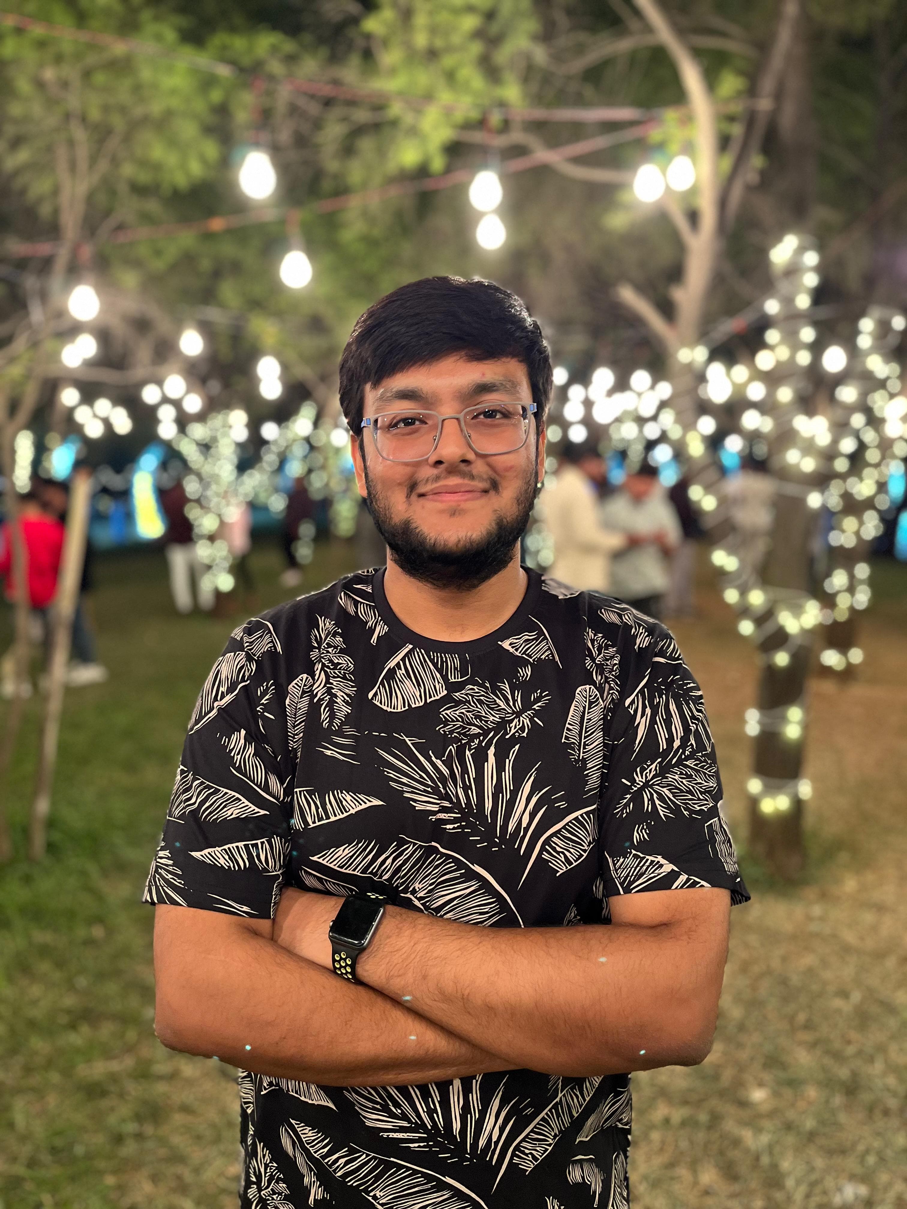 Anchit Shrivastava outdoors on a warm evening in a garden, arms crossed and smiling, wearing glasses and a black patterned t-shirt, with soft string-light bokeh glowing behind him.